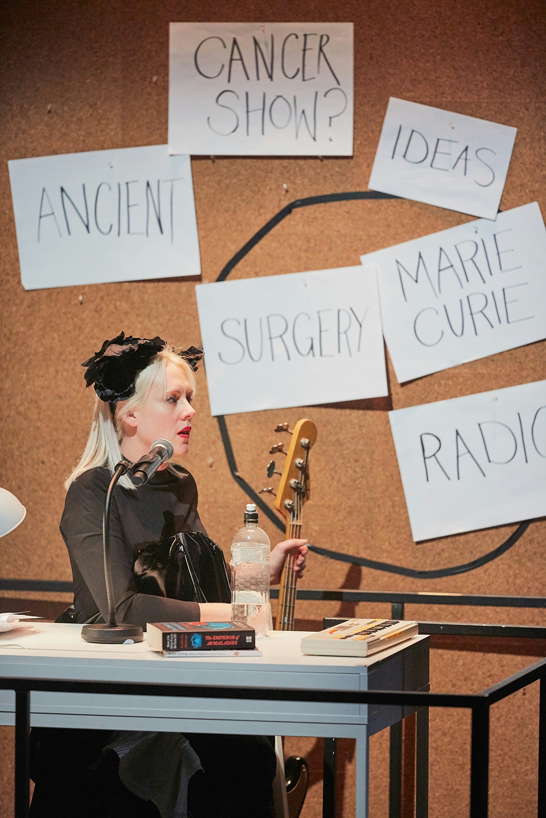 A woman with a guitar sits at a desk. There’s a noticeboard behind her with words pinned up behind her: ‘Cancer show?’, ‘Ancient’, ‘Ideas’,  ‘Surgery’, ‘Marie Curie’.