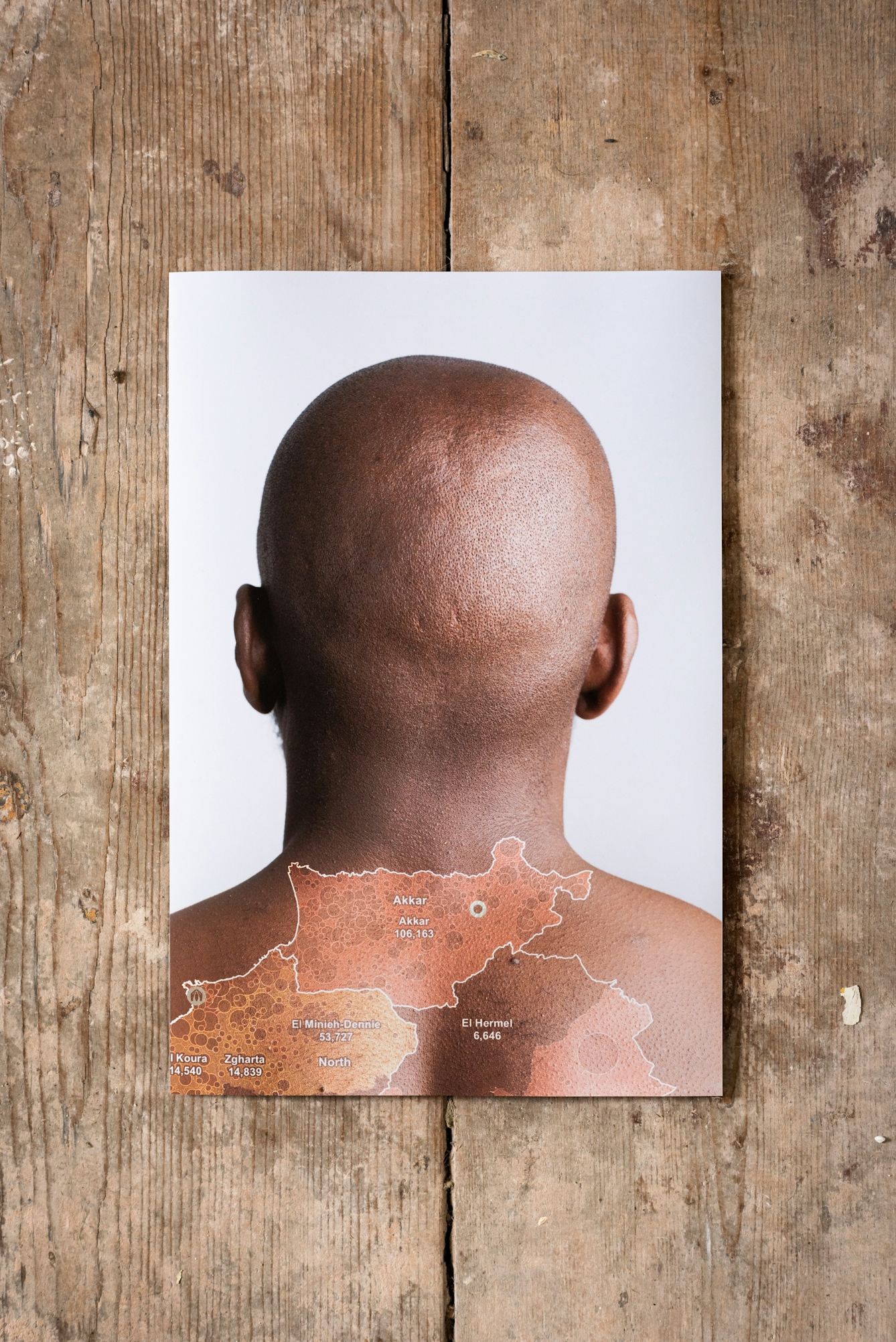 Photograph of a photographic print on a weathered, paint spattered wooden floor. The print shows the naked back of a man's head and neck against a white background. Superimposed on his back is a simple line map in white, yellow and orange, outlining different regions.