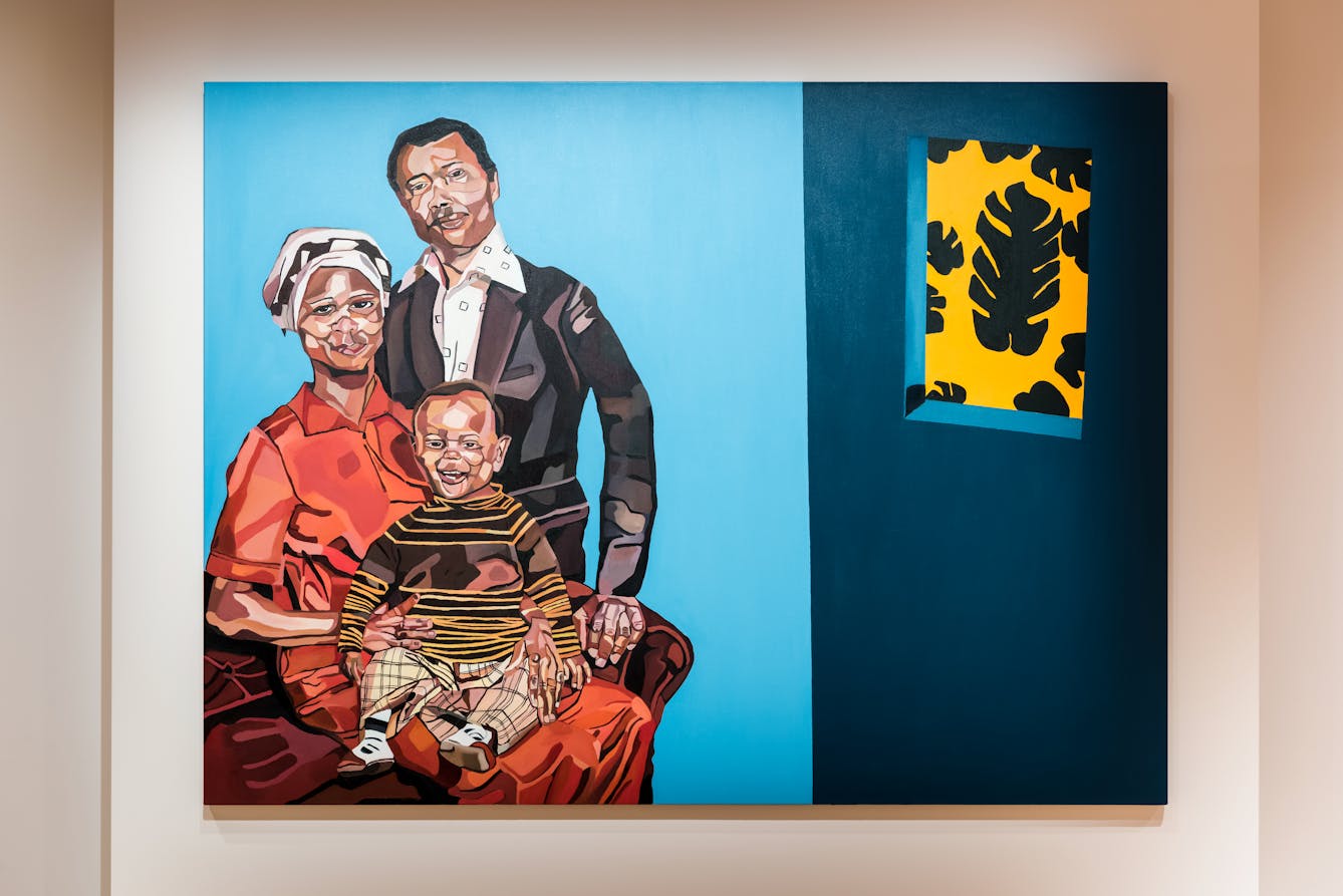 Photograph of an exhibition gallery space showing a section of light pink coloured wall on which a large colourful oil painting has been hung. The painting shows a family portrait of a seated mother with a young child on her lap and a standing father figure. The background of the painting is shades of blue with a window out to a yellow background with black leaf motifs.