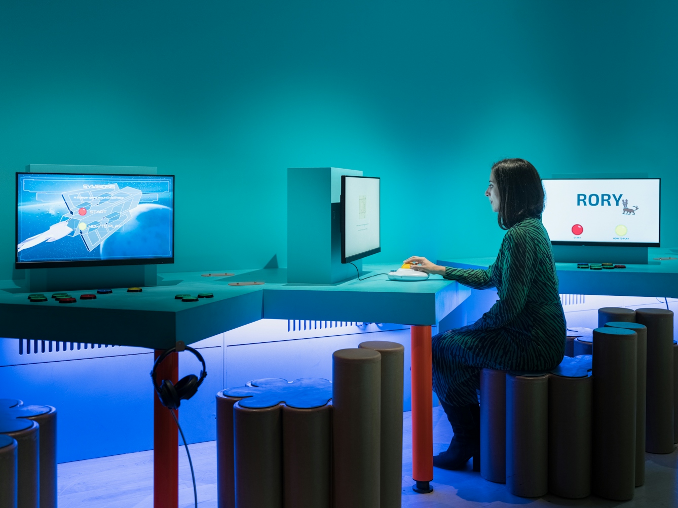 Photograph of a woman in an exhibition space seated at a computer monitor with her hand on a large track ball. To her left and right are two other screens with buttons on the table top in front of them.