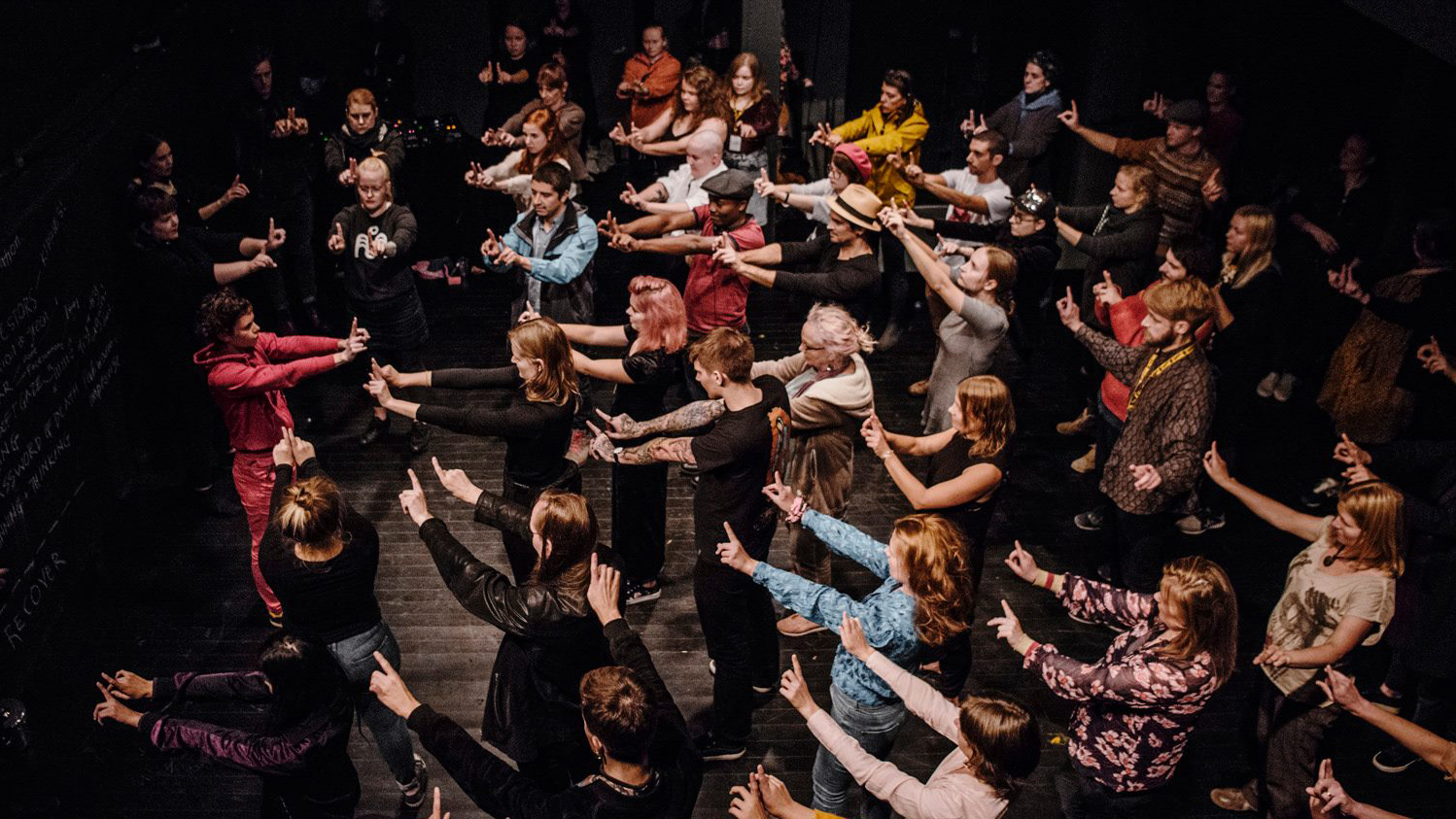 A photograph taken from above of people taking part in a group activity. They are facing the same direction and holding both arms out in front of them with their index fingers pointing.