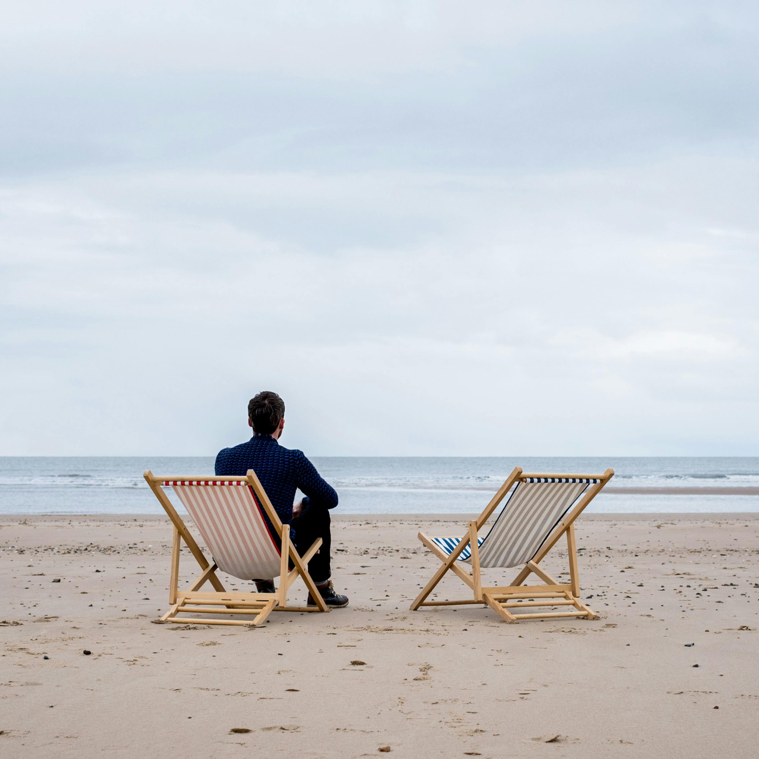 Photograph of a young man sat in a deck chair on a sandy beach, looking out to sea. To his right is an empty deck chair.