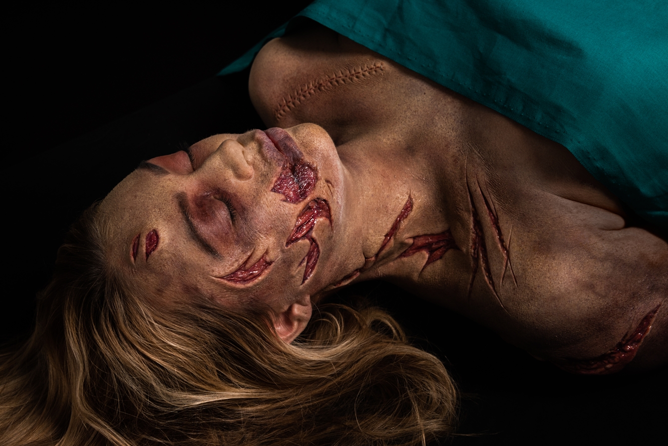 Photograph of a the head and shoulders of a woman lying on her back on a black background. Her chest is covered with a green surgical sheet. She has her eyes closed and her blond hair falls back onto the tabletop. She has prosthetic makeup applied which creates the realistic effect of her face, neck and shoulders having been slashed with a knife. The makeup reveals the layers below the skin. By her left shoulder there is makeup which creates the impression of a long surgical cut which has been stitched up.