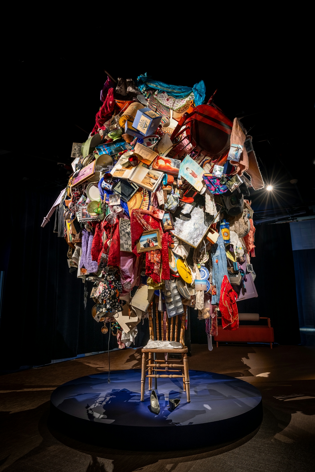 Photograph of an exhibition installation showing a solitary chair placed in the centre of a blue circular plinth. Under the chair are a pair of silver stilettos. Above the chair, a multitude of objects seem to be falling out down from the dark ceiling. These objects include clothes, photo albums and furniture.