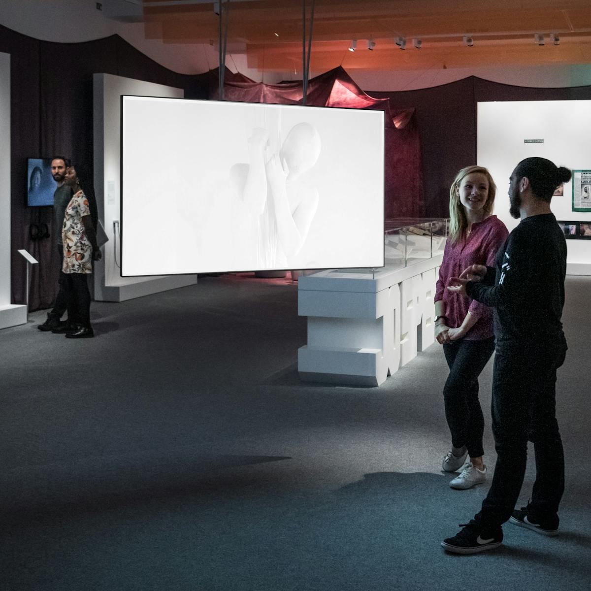 Photograph of the Misbehaving Bodies exhibition at Wellcome Collection, showing a wide view of the space. in the foreground a young man and woman discuss a video piece projected onto a large screen. In the background an other young man and woman look at the artworks that are displayed on the wall.