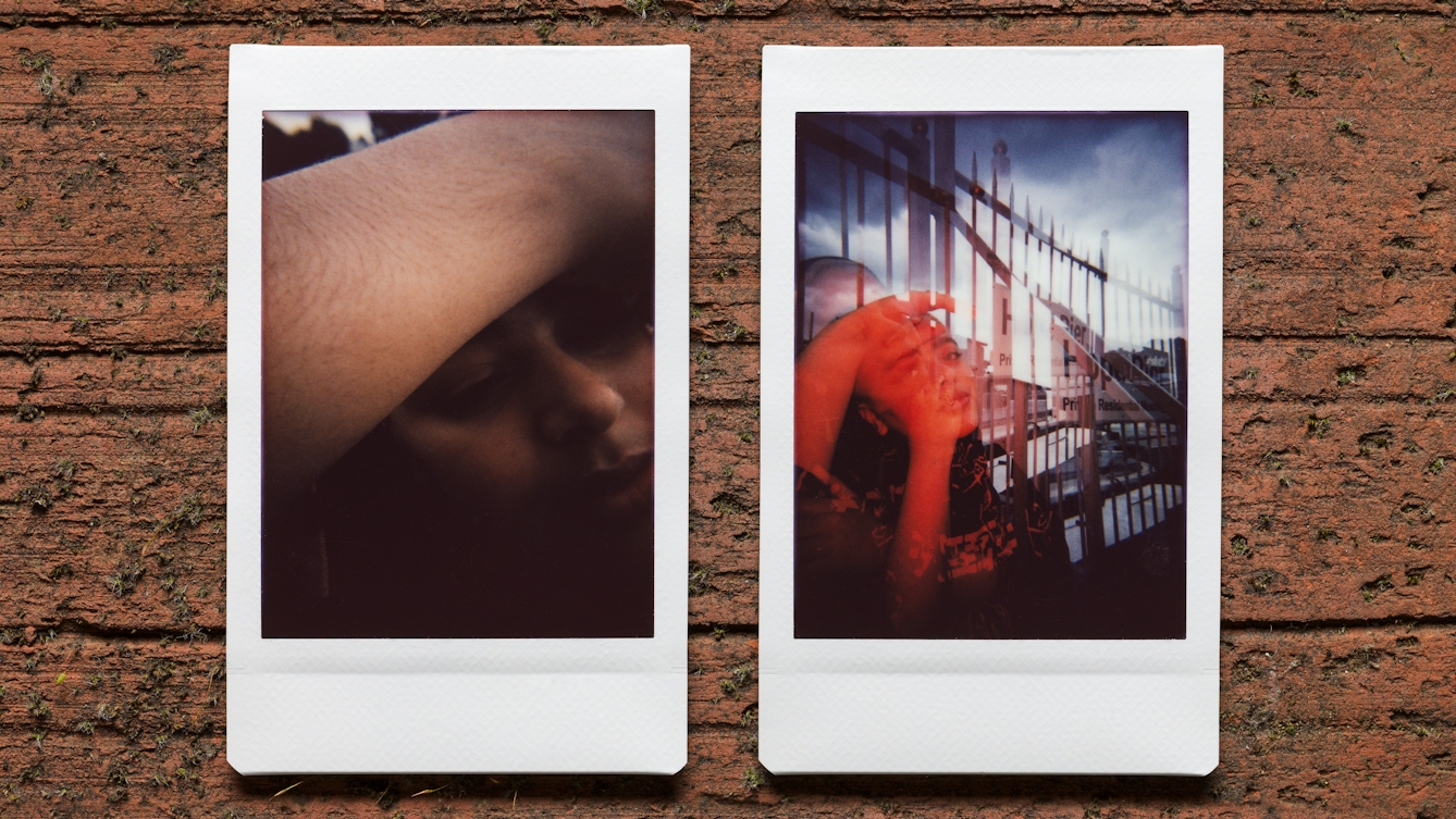 Photograph of two Instax Mini instant film prints in a line, resting on a textured brick surface. The two prints feature the same woman. The print on the left shows a close-up of her face, with her right arm raised over their forehead showing the hair on her arm. The print on the right shows the her reclining against a set of railings in an outside urban scene. Her face is lit with a red light and there is a sense of a double exposure.