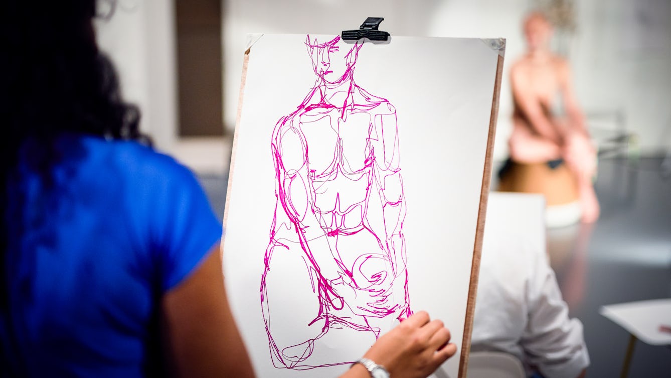 Photograph of a life drawing workshop showing a young woman sketching at a charcoal drawing of a male figure who can be seen posing in the background.