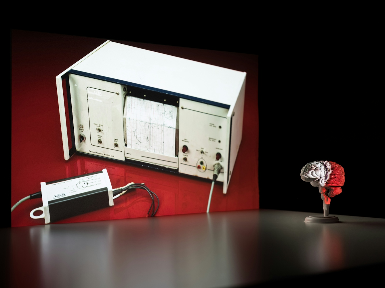 Photograph of a dark corner of a room. On the wall is a projected photograph of a red table on which is a large white scientific monitoring machine, complete with dials and a paper readout. In front of the projected image is a flat tabletop which shows a slight reflection of the illustration. Standing on the table to the right is a small model of a brain, resting on a stand. Part of the projected image washes across the surface of the brain, covering it in the image and its colours.