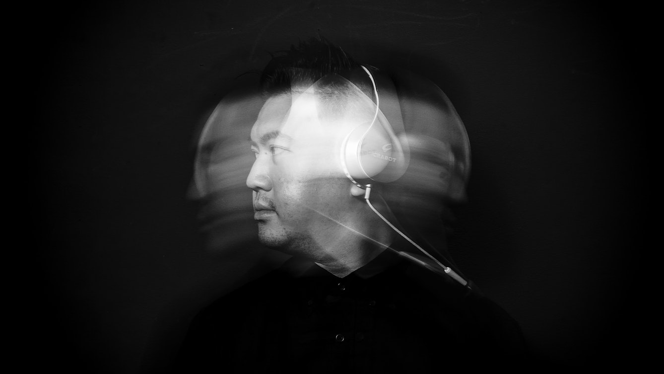 Black and white photographic portrait of a man dressed in a black shirt wearing a pair of over ear headphone with the white cables trailing over his shoulder. He is pictured from the chest up. The man's face is spotlit in a small circle of light. He is standing against a black background which means he is surrounded by darkness. He is moving his head from side to side causing his head to motion blur in an arc across the image. A flash light has caught this head as it is turned to the left, creating a sharp image within the blur.