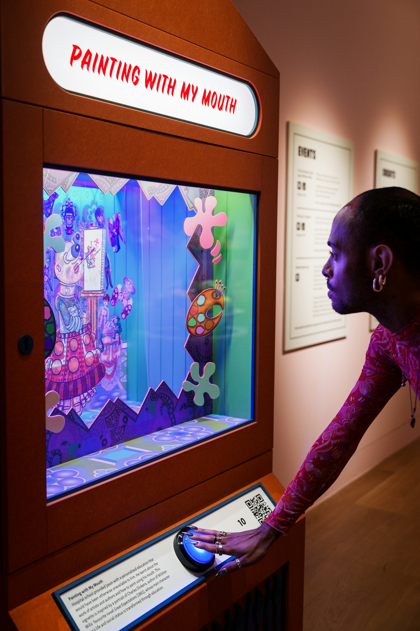 Photograph of a gallery visitor activating a large button on the front of an arcade style gaming machine. They are looking into a large glass fronted diorama, inside which is a multi-layered theatre style scene containing colourful drawings of characters and scenery.The backlit title of the diorama appears at the top of the booth and says 'Painting with my mouth'.