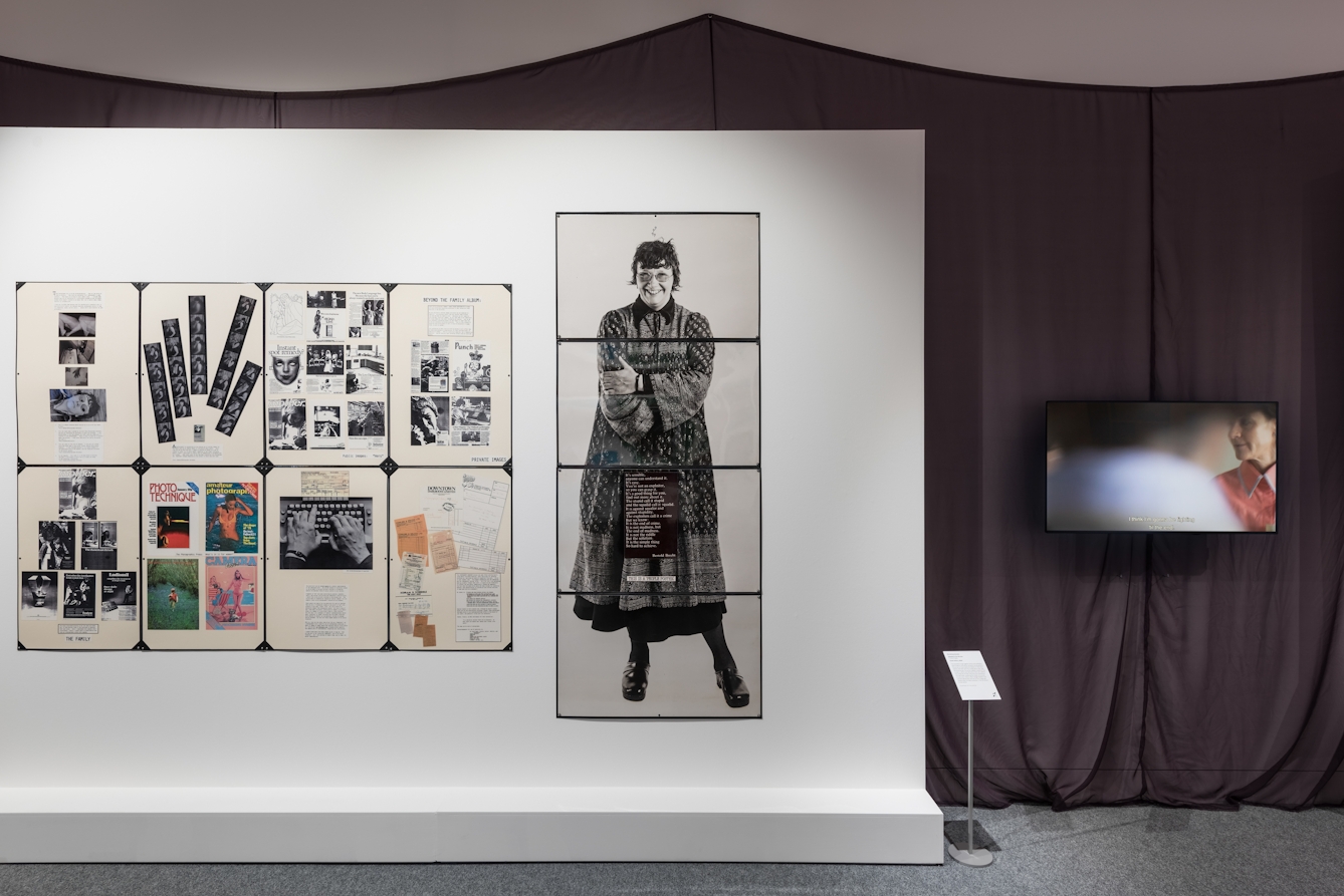 Photograph of a gallery wall showing several laminated sheets of collaged text and photographs. Behind the gallery wall is a draped fabric. to the right is a television screen showing a still from a film.