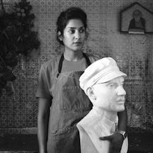 Black and white photograph of a young woman with dark hair wearing an apron; she stands behind the bust of a white statue, with one arm over its shoulder. In the background a plant and picture can be seen on the wall.