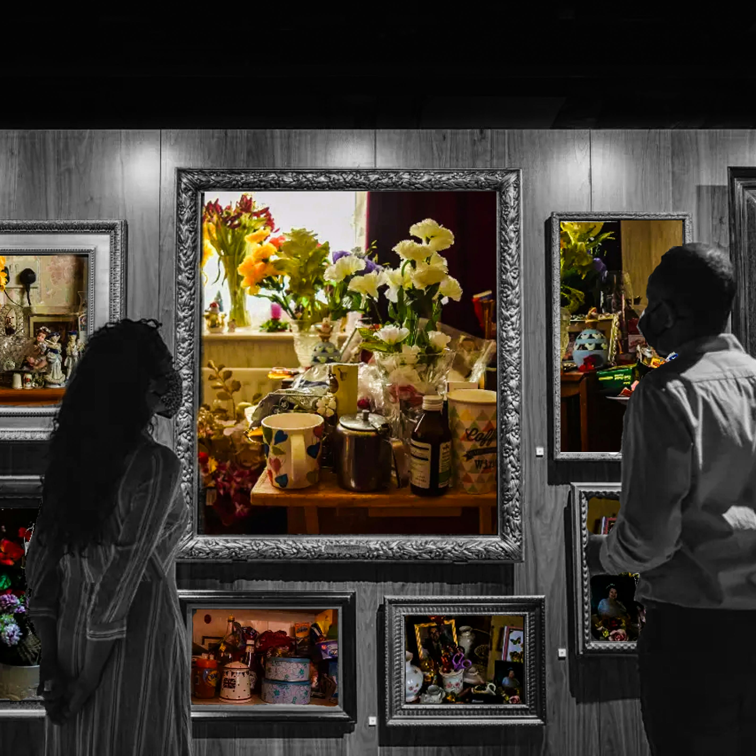 Black and white photograph showing a wall in a museum covered in large ornate picture frames. In the foreground are two people facing the wall of frames and wearing masks. Inside the black and white frames are colourful photographs showing different household objects. The largest frame in the center shows a table that has lots of things on top of it, all packed very close together, including vases of flowers, a mug and coffeepot, and medicine. On the windowsill behind the table are more flowers. The frame to the right shows a photo of a an equally crowded table, holding photo frames, flowers and various other trinkets. 