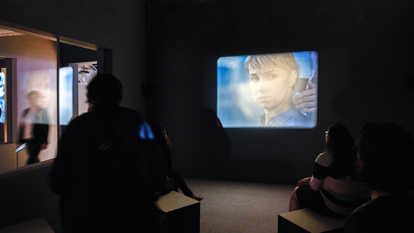 Photograph of film projection showing a woman's face, within a dark gallery setting. In the foreground are visitors sitting on benches watching the film.