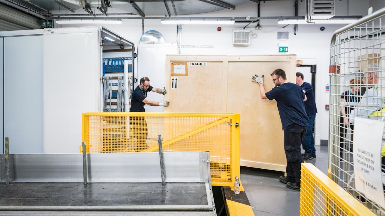 Photograph of a loading bay showing a white panel van with its back doors wide open and tail lift resting on the raised concrete floor. Three men in blue and black work clothes are wheeling a large wooden crate into the back of the van. On the side of the crate are 2 black arrows pointing toward the top and with word 'FRAGILE'.