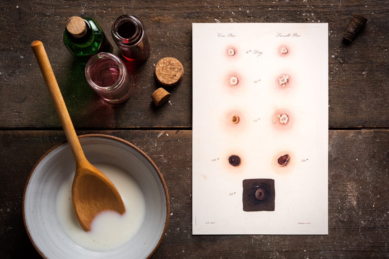 Photograph of a weathered wooden tabletop. Laying flat on the table is a sheet of paper with close up illustrations of the various of stages of cow-pox and smallpox. Also on the tabletop are glass bottles containing coloured liquid with cork stoppers and a bowl containing a white liquid with a wooden spoon resting inside.