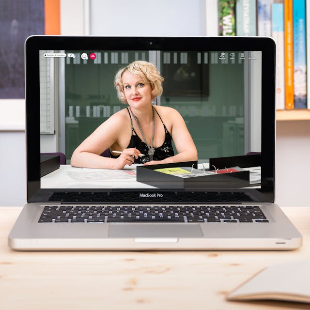 Photograph of a laptop on a desk, on the screen is a photographic portrait of Dr Kate Lister sat at a table with a selection of archive material from Wellcome Collection’s sex worker card collection laid out in front of her. Around the laptop is an open notebook and pen, a pile of books and pens in a pot. In the background are bookshelves. 