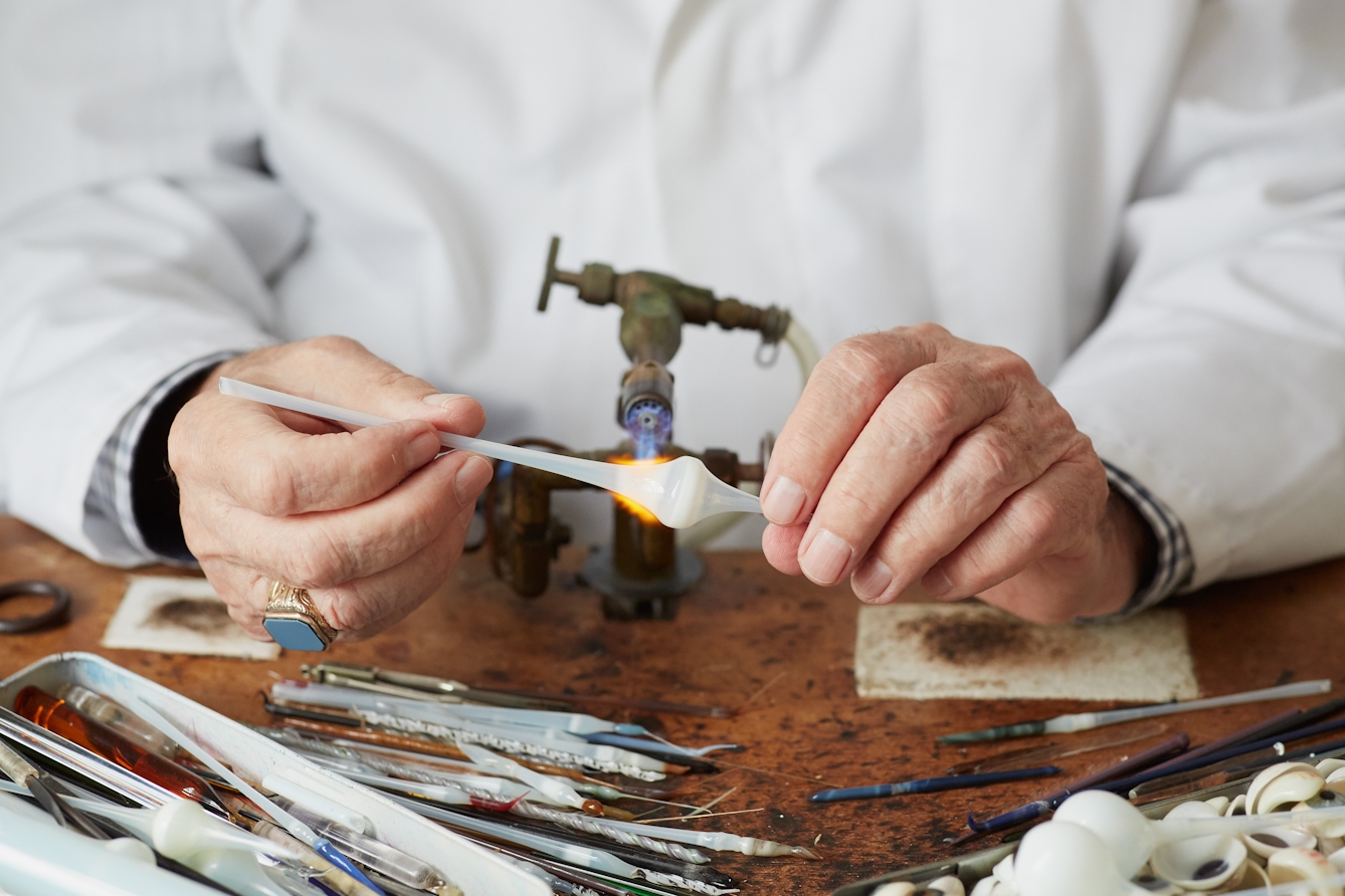 Photograph of the hands of a man in a white lab coat holding a length of white glass tubing to a gas burning torch. The tube is expanding where it meets the heat of the flame. On the workbench in front of him are think tools and other lengths of glass tubing. 