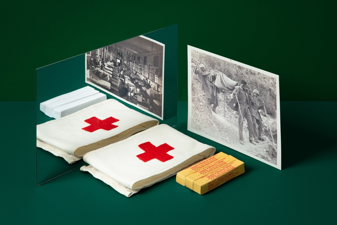 A photograph of a mirror on a dark green background. In the foreground is a medics armband with a Red Cross, packages of painkillers and a photograph of field medics from the Second World War. In the reflection of the mirror is a different photograph featuring a filed hospital and blank white painkiller packages.