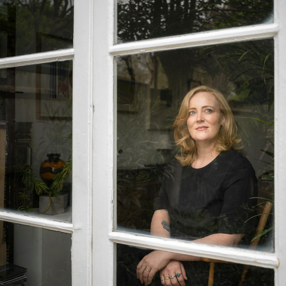 Photograph of a blonde woman with black clothing looking through a window, captured from outside.  The woman is sitting on a wooden chair with her arms crossed at the wrists with her gaze towards the top right of the frame.