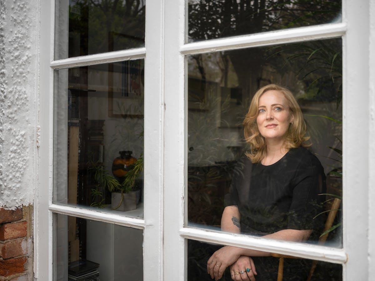 Photograph of a blonde woman with black clothing looking through a window, captured from outside.  The woman is sitting on a wooden chair with her arms crossed at the wrists with her gaze towards the top right of the frame.