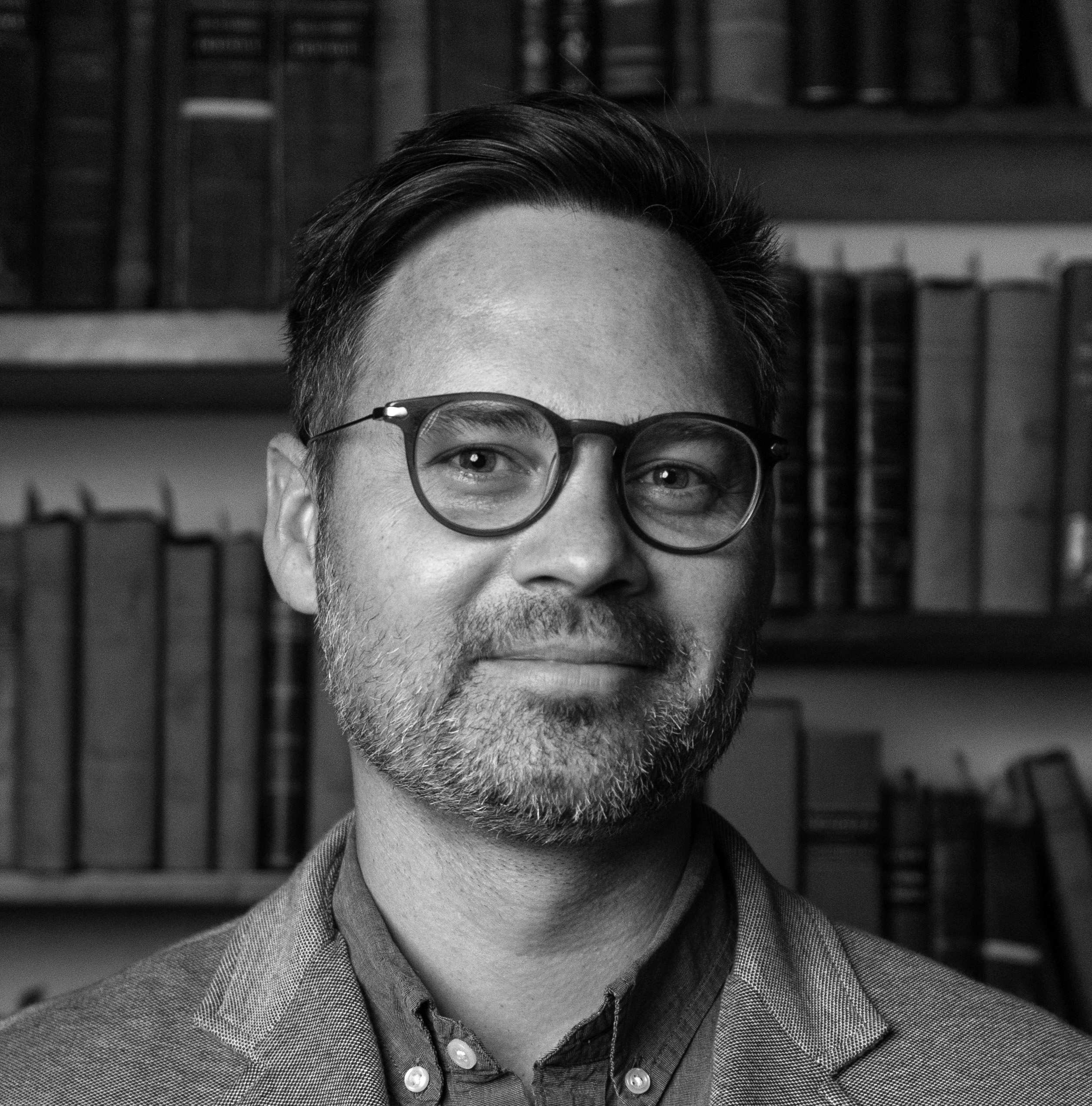 Black and white photograph of a white man with round, plastic framed glasses standing infront of bookshelves and smiling directly at the camera.