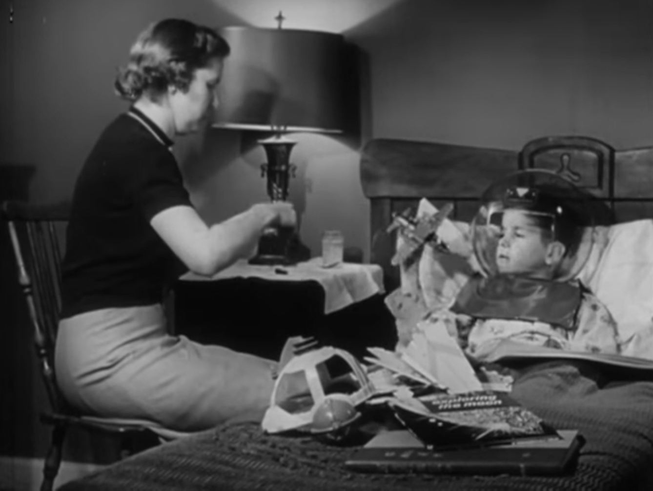 Still from black and white film featuring a boy in bed wearing an astronaut helmet costume, and a woman sat on a chair next to his bed.