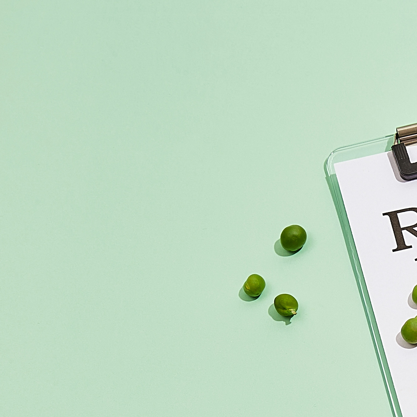 Photograph of a clipboard with a blank doctor's prescription note attached to it, resting on a light green background. Loose on the background, to the left of the clipboard are a scattering of loose green peas.
