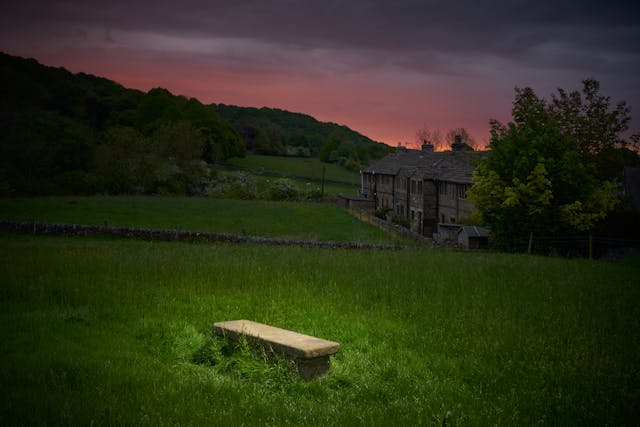 Photograph of a rural landscape at dusk. In the background is a row of houses. In the foreground is a spotlit plain stone grave.