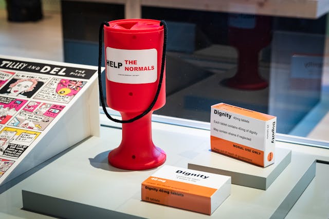 Photograph of an exhibition display case, which contains a red collection box bearing a sticker which says, 