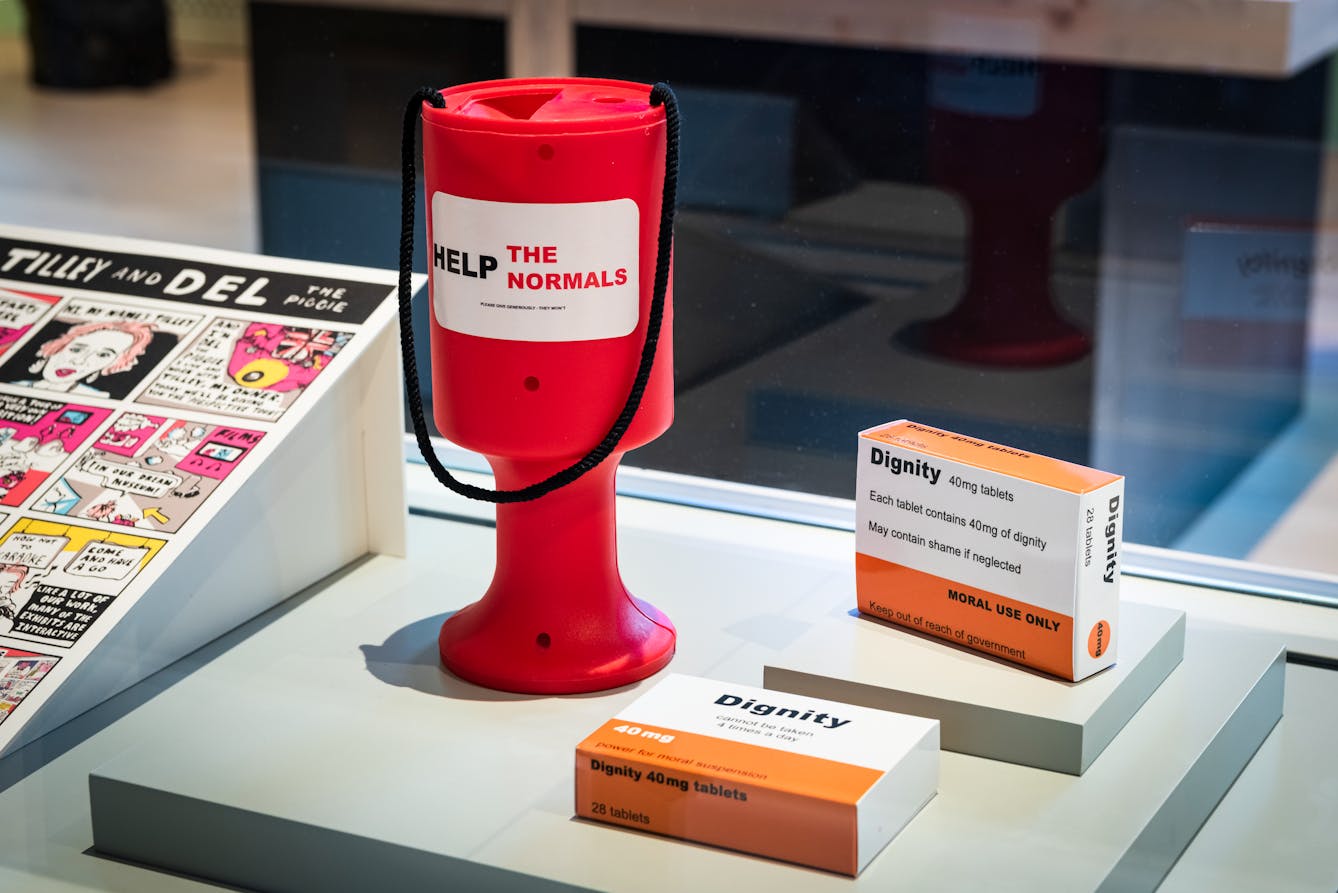 Photograph of an exhibition display case, which contains a red collection box bearing a sticker which says, 'Help the normals'. Next to the collection box on one side is part of a comic strip and on the other side are 2 pill boxes with the words 'Dignity' and other smaller text printed on the packaging.
