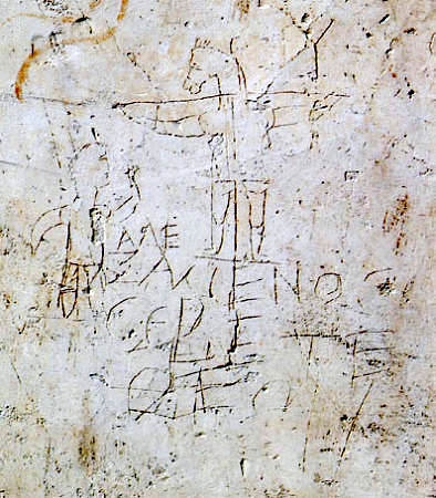 Photograph of an image and words etched into a stone material. The sketch shows a crucified man with the head of a donkey and another man looking on. The text tells us “Alexamenos worships his god.”