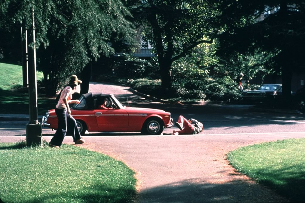 Image of a stationary red car in a road with a man lying on the pavement in front of it. Two onlookers are close by.