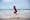 Photograph of a young child with short blonde hair jumping into the air by the sea on a sandy beach. They are wearing a blue swimming top and a pair of red shorts. As they jump they are holding on to a red brimmed hat on their head
Further into the distance is the horizon line of the sea. The sky is blue and cloudy.