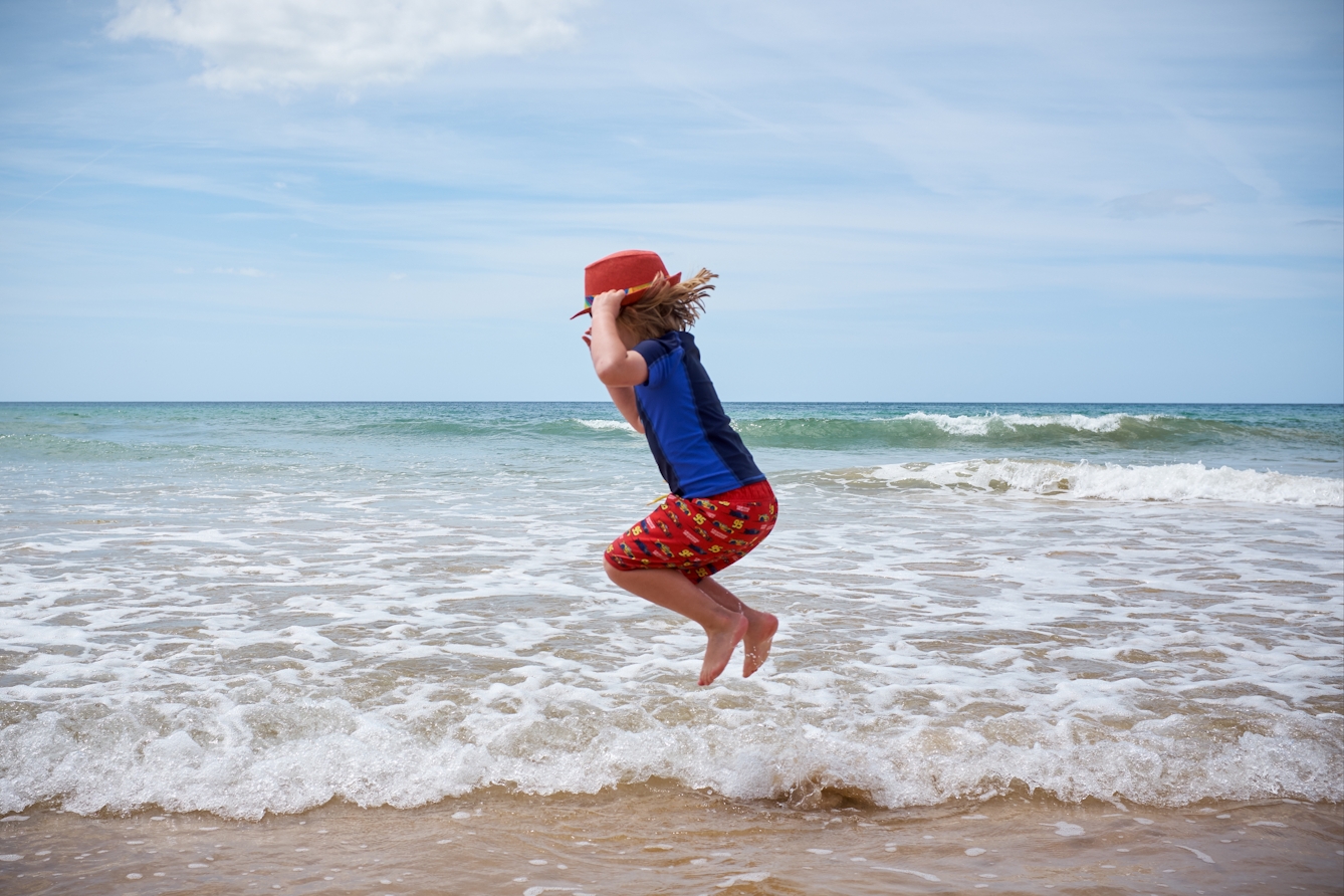 Photograph of a young child with short blonde hair jumping into the air by the sea on a sandy beach. They are wearing a blue swimming top and a pair of red shorts. As they jump they are holding on to a red brimmed hat on their head 

Further into the distance is the horizon line of the sea. The sky is blue and cloudy. 