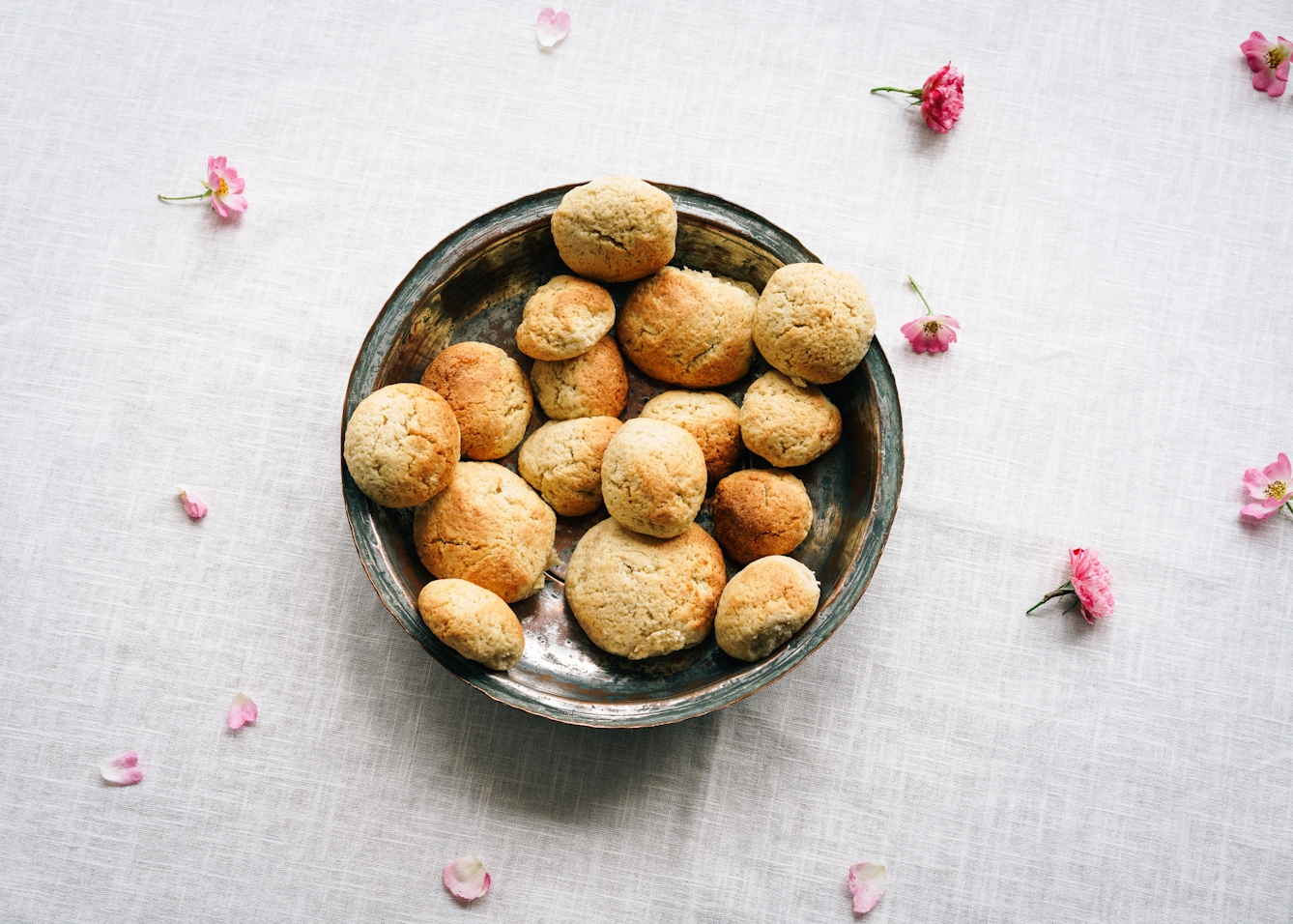 Photograph of 16 golden backed balls made of a sort of dough, piled in a dark ceramic bowl, viewed from above. The bowl stands on a white linen tablecloth, which is scattered in flower heads and petals.