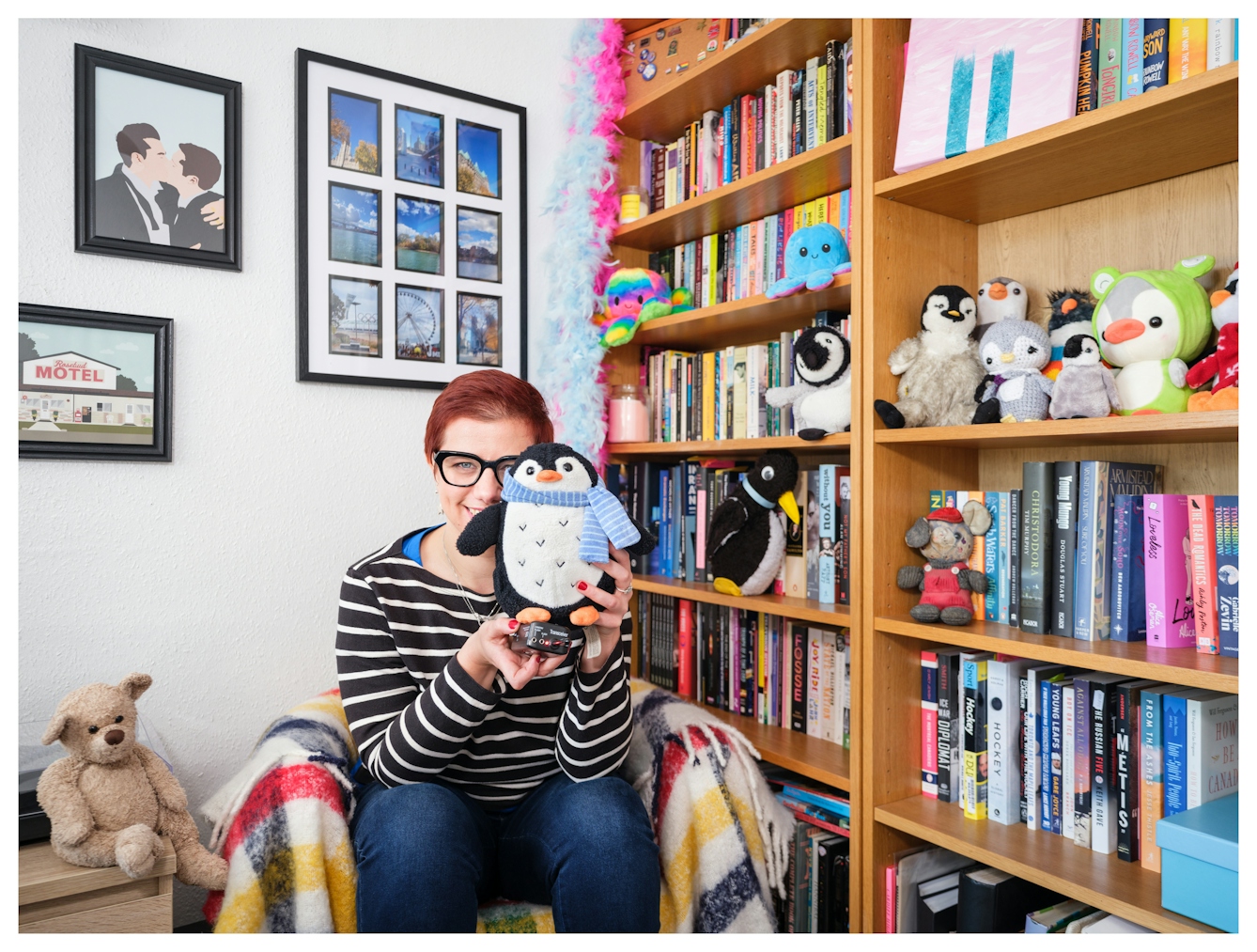 Assisted Self-Portrait of a woman sat leaning forward in a low armchair in a domestic setting. To the right are large book cases filled with books and dotted with soft toy animals. She is hiding a small soft toy penguin up to her face, obscuring the left hand side. She is smiling broadly and hold the remote trigger in her right hand that she used to capture the image.