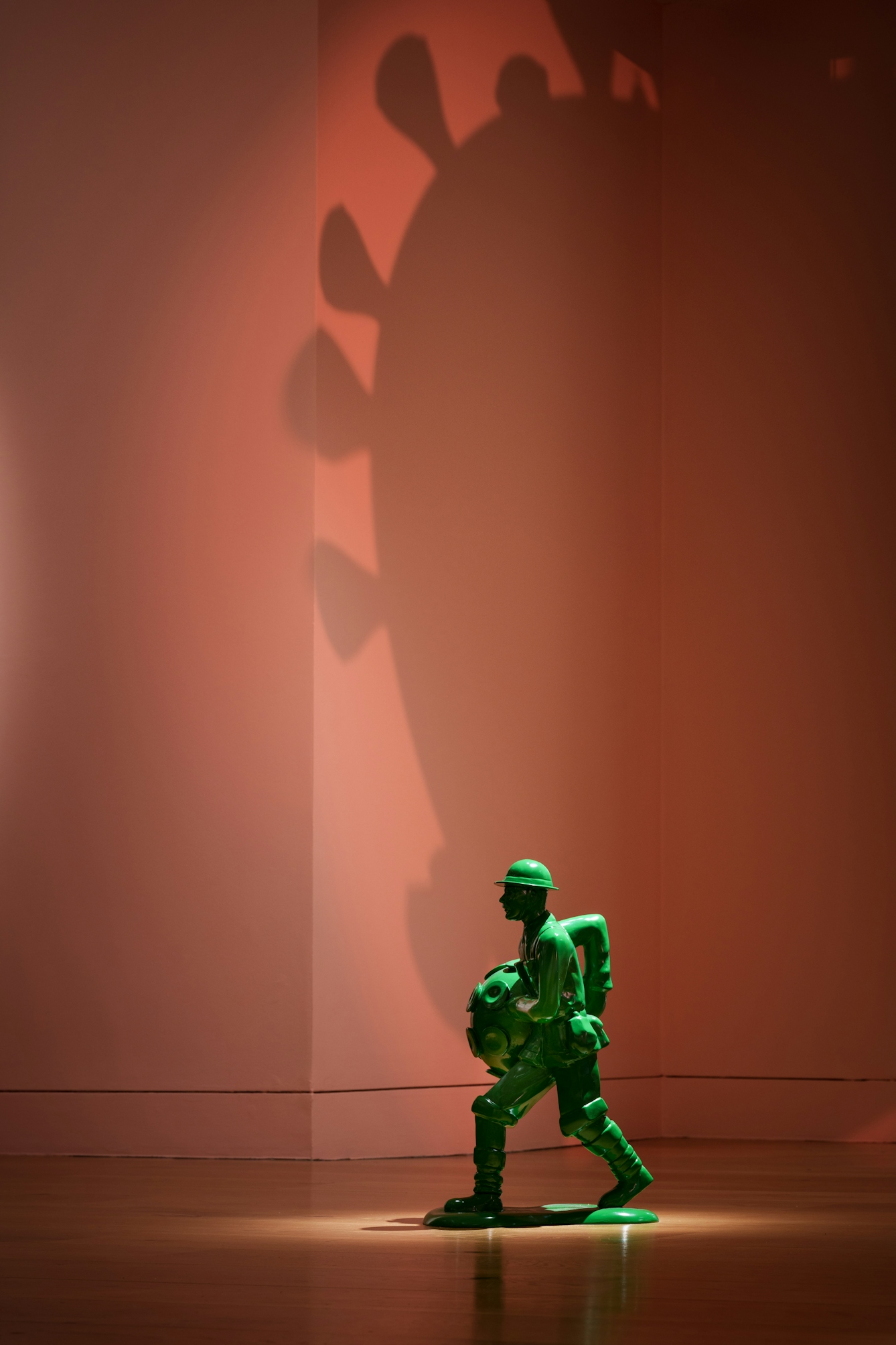 Photograph of an oversized green toy soldier sculpture wielding a molecular representation of a virus. Behind the soldier a large shadow of the virus is cast on the wall.