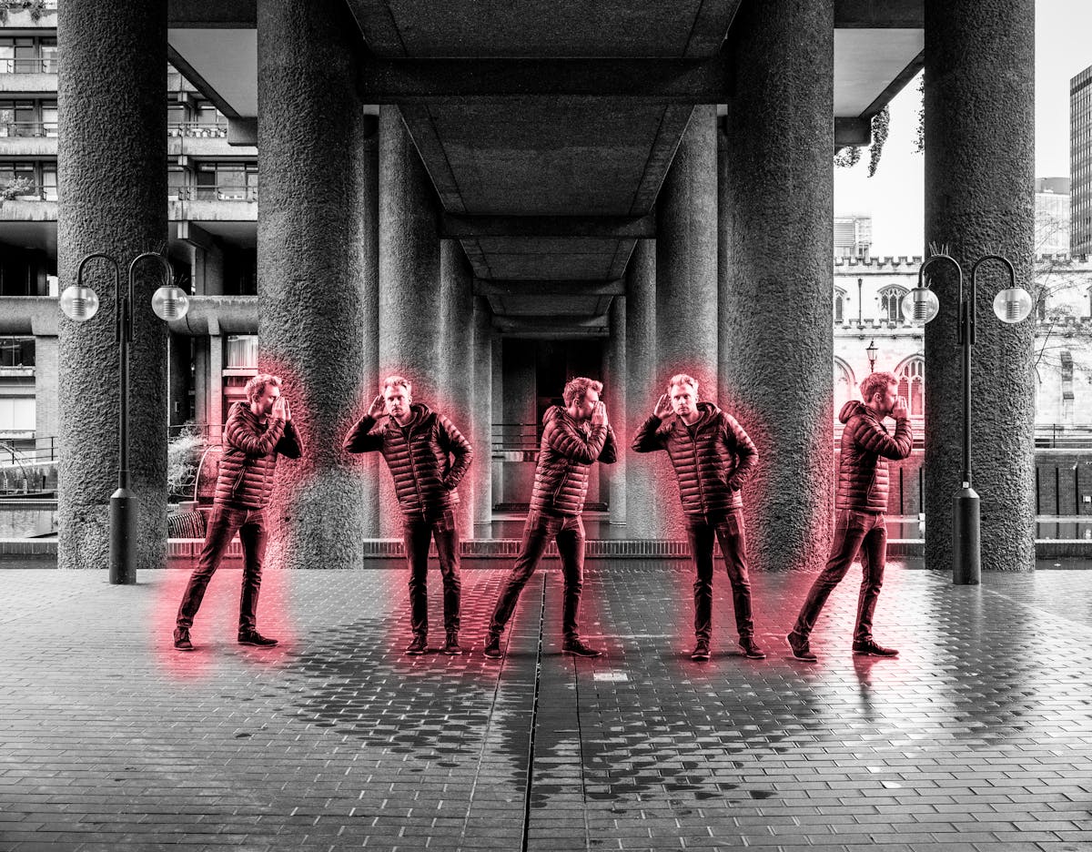 Black and white photograph of a concrete urban environment with an image of the same man repeated 5 times across the image. In each instance he is highlighted by a red glow. He appears to be passing a verbal message along to line to himself.