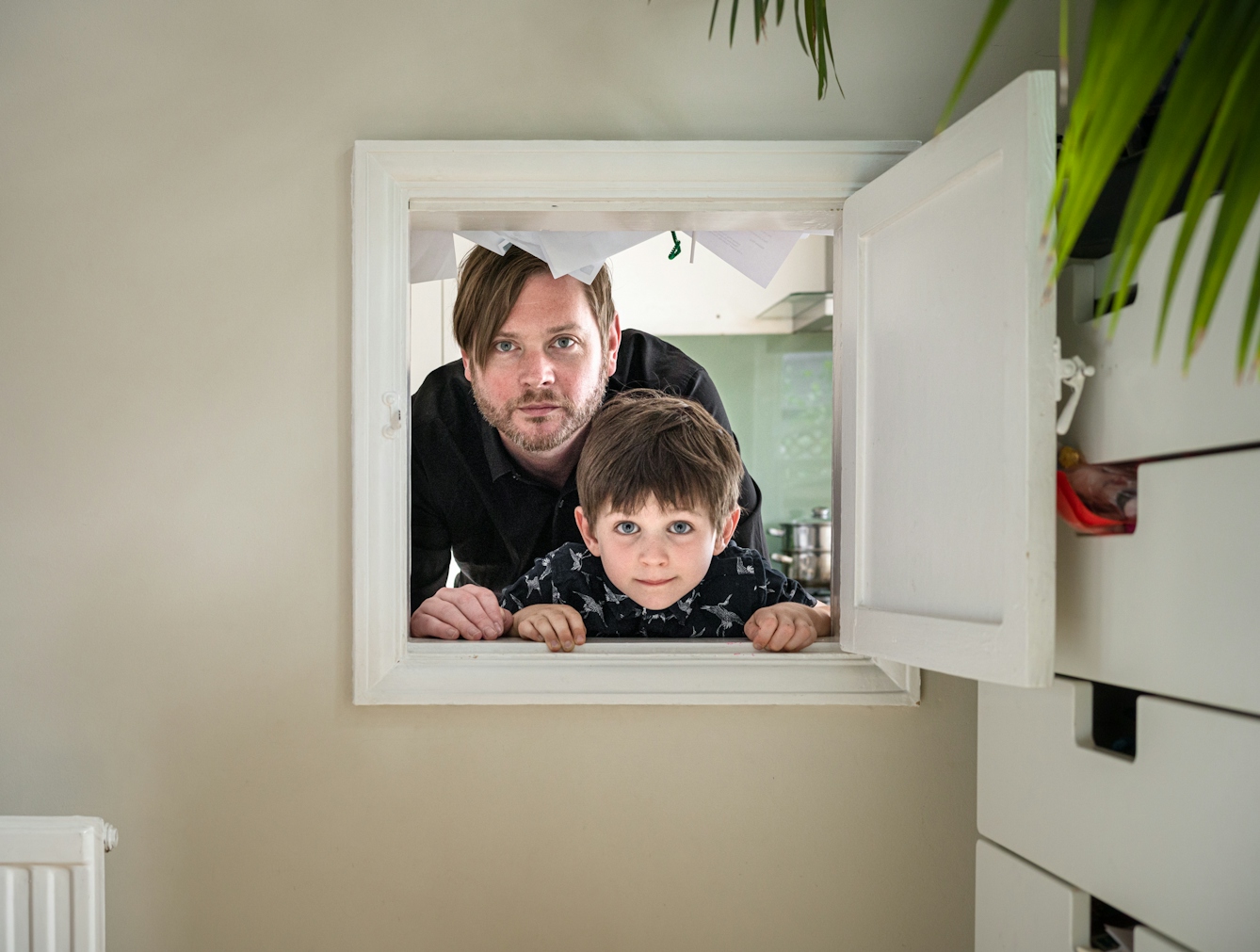 Photograph of a man and his son looking through a kitchen hatch. To the right is a chest of drawers. 
