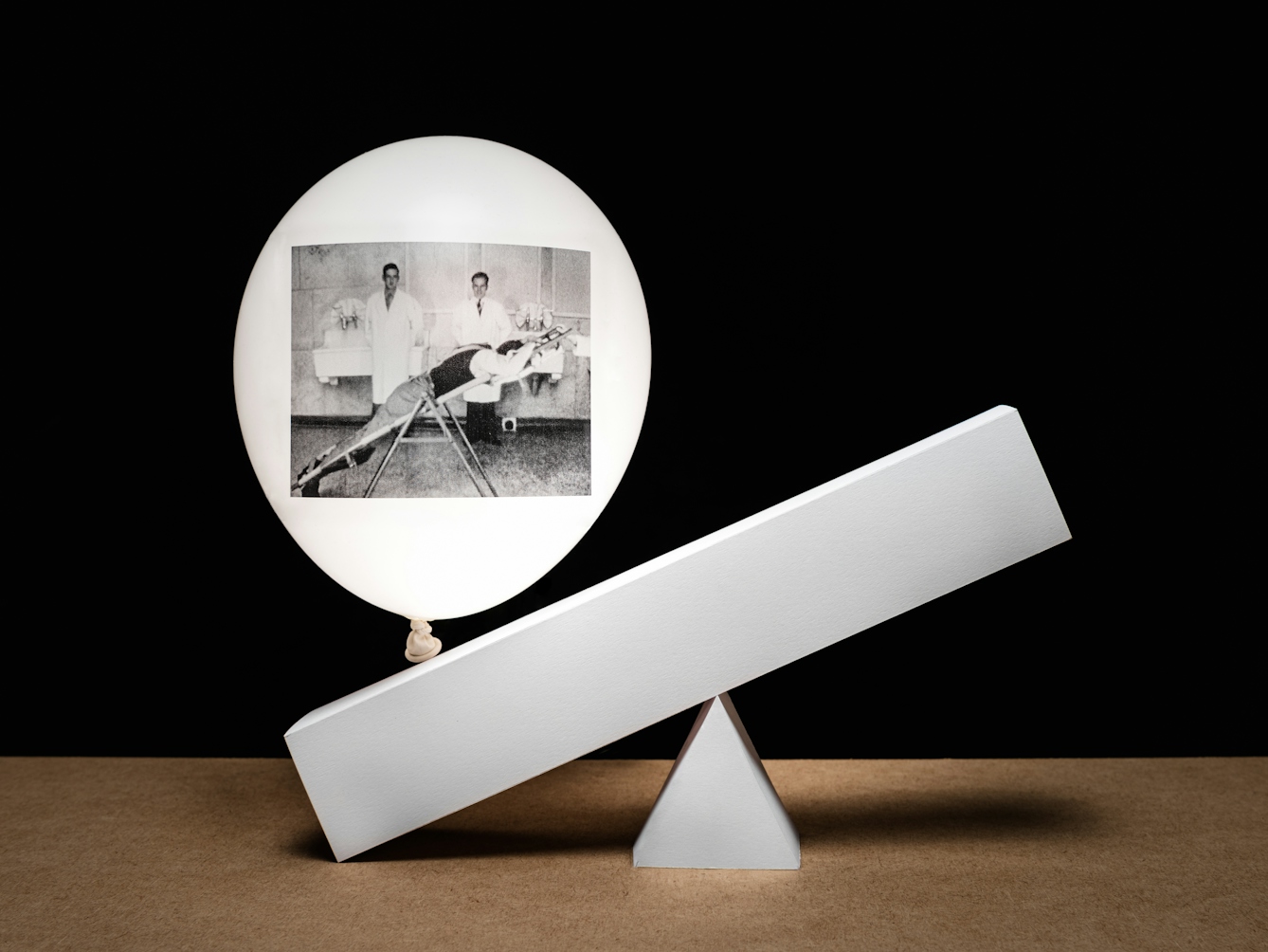 Photograph of a white inflated balloon against a black background, floating vertically above a wooden tabletop horizon line. The balloon looks like it is illuminated from within. On the side of the balloon is a rectangular, monochrome archive film still. The still shows a man lying prone on a pivoted flat board. Being him are standing two men in white medical coats. The balloon is floating above two three dimensional graphic elements made of white card. One is a triangle on which is resting a long rectangle, sloping down to the tabletop at the left hand end. The shape of these 3D elements echo the pivot contraption shown in the film still.