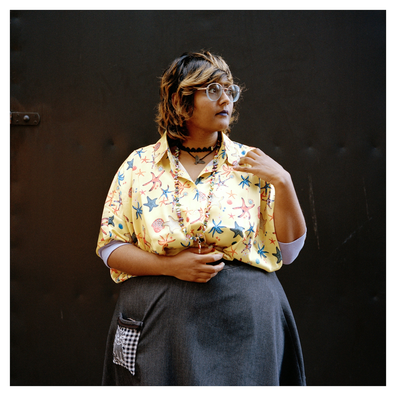 Portrait photograph of Sumita, a woman with shoulder length brown hair with blonde streaks. She is wearing a yellow shirt with a starfish pattern and a grey skirt. She is wearing glasses, purple lipstick and several chunky necklaces. 

She is standing in front of a brown backdrop and is looking to her left, with a neutral expression.