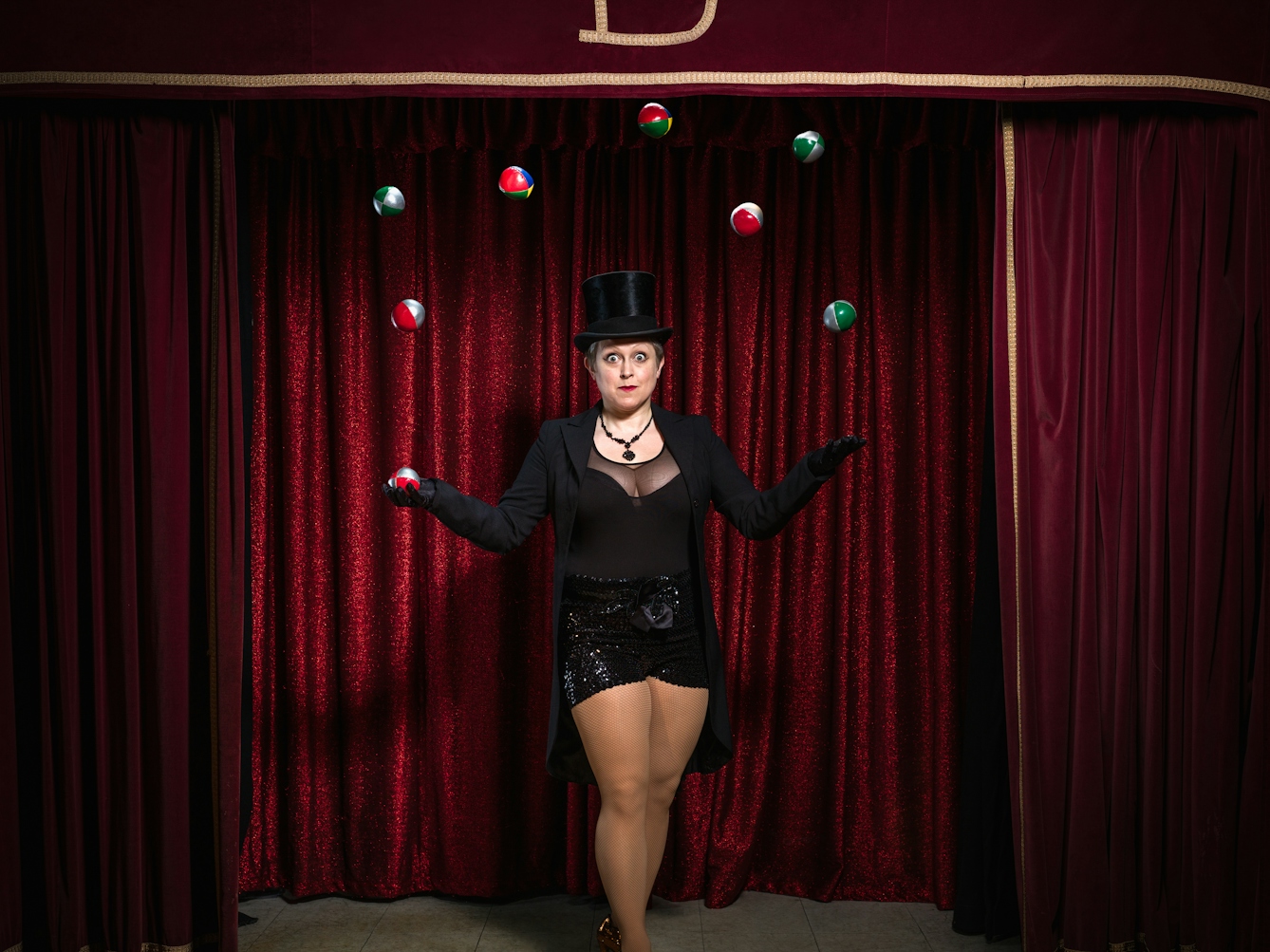 Photograph of a woman on a theatre stage surrounded by red curtains, dressed in a top hat and tails juggling 8 balls in the air.