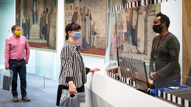 Photograph of the entrance desk of a library. Behind the desk a man stands wearing a face covering and a staff lanyard. He is talking to a female visitor standing on the other side of the desk, who is also wearing a face covering, a stripy jacket and who is holding a plastic bag. Between the two of them is a large perspex screen which runs the length of the desk. Queuing behind the visitor at the desk is a man in a pink shirt and a yellow face covering who is holding a see through plastic bag containing a laptop. On the glass partition in the background are two large figurative oil paintings.
