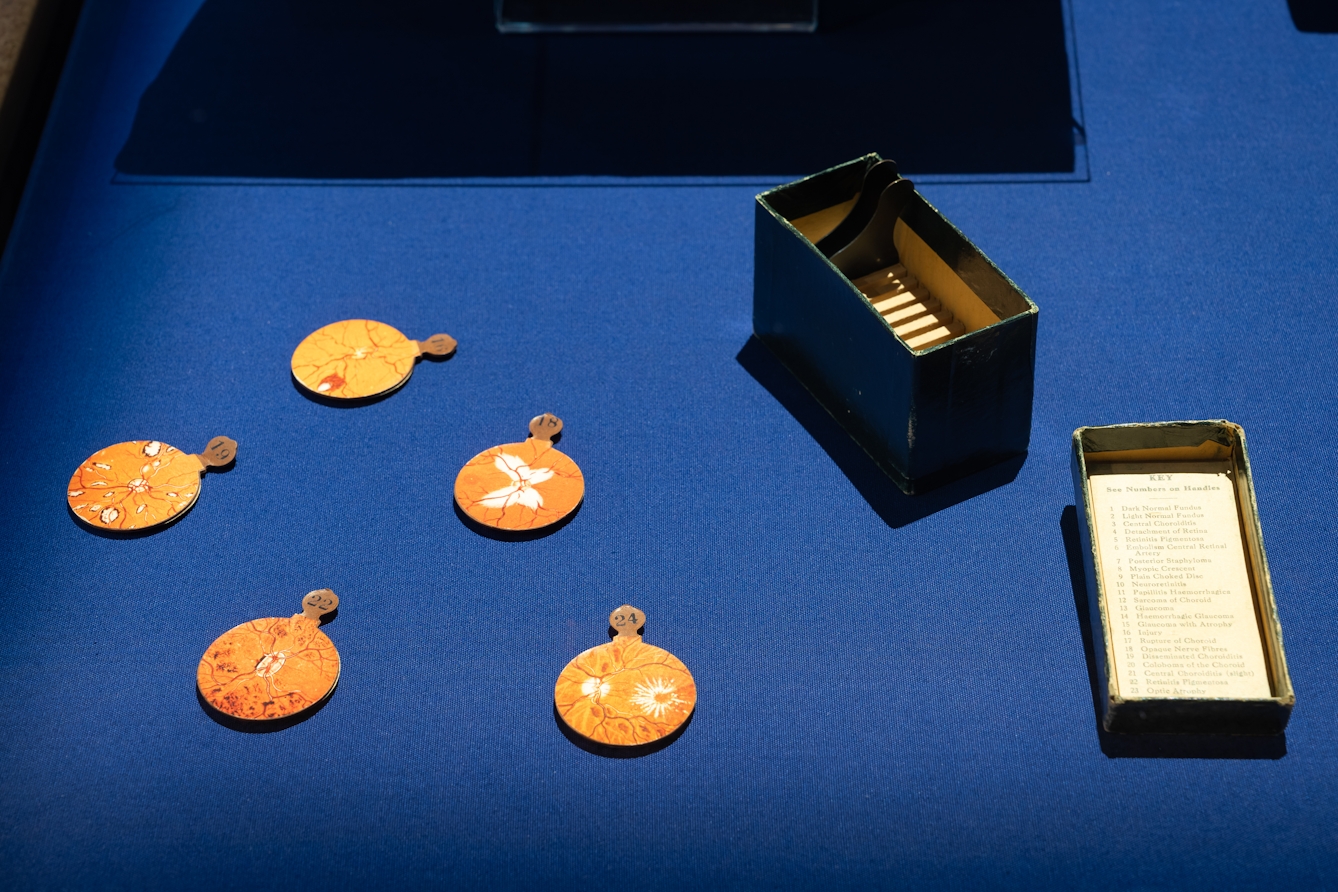 Photograph of blue fabric base to an exhibition display case. On the base are 5 numbered discs each with a diagrammatical drawing in oranges, whites and thin squiggly red lines. Next to them is the open box in which they were stored. The lid has a typed sheet of paper which describes the content of each disc.
