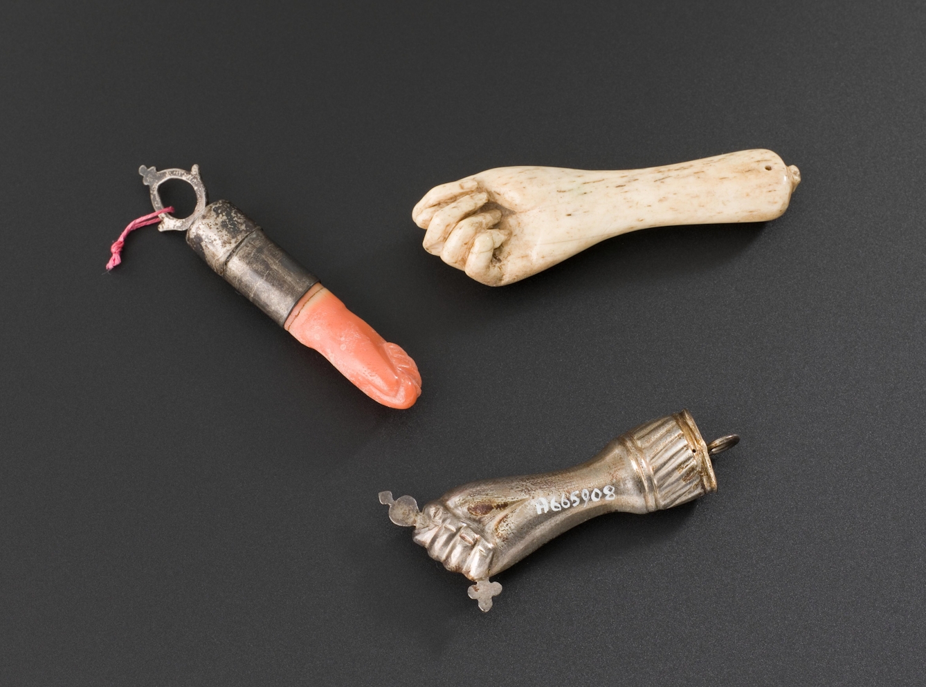 Photograph of a set of 3 amulets on a textured grey background. All three represent the forearm and hand of a human, made out of different materials including bone and metal.