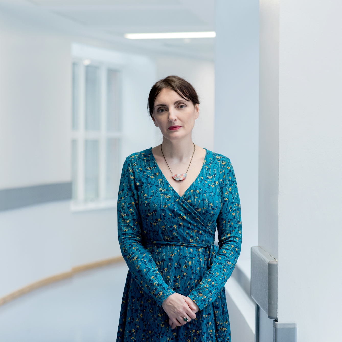 Photographic portrait of a woman wearing a blue patterned dress looking straight to camera, standing in a plain white hospital corridor.