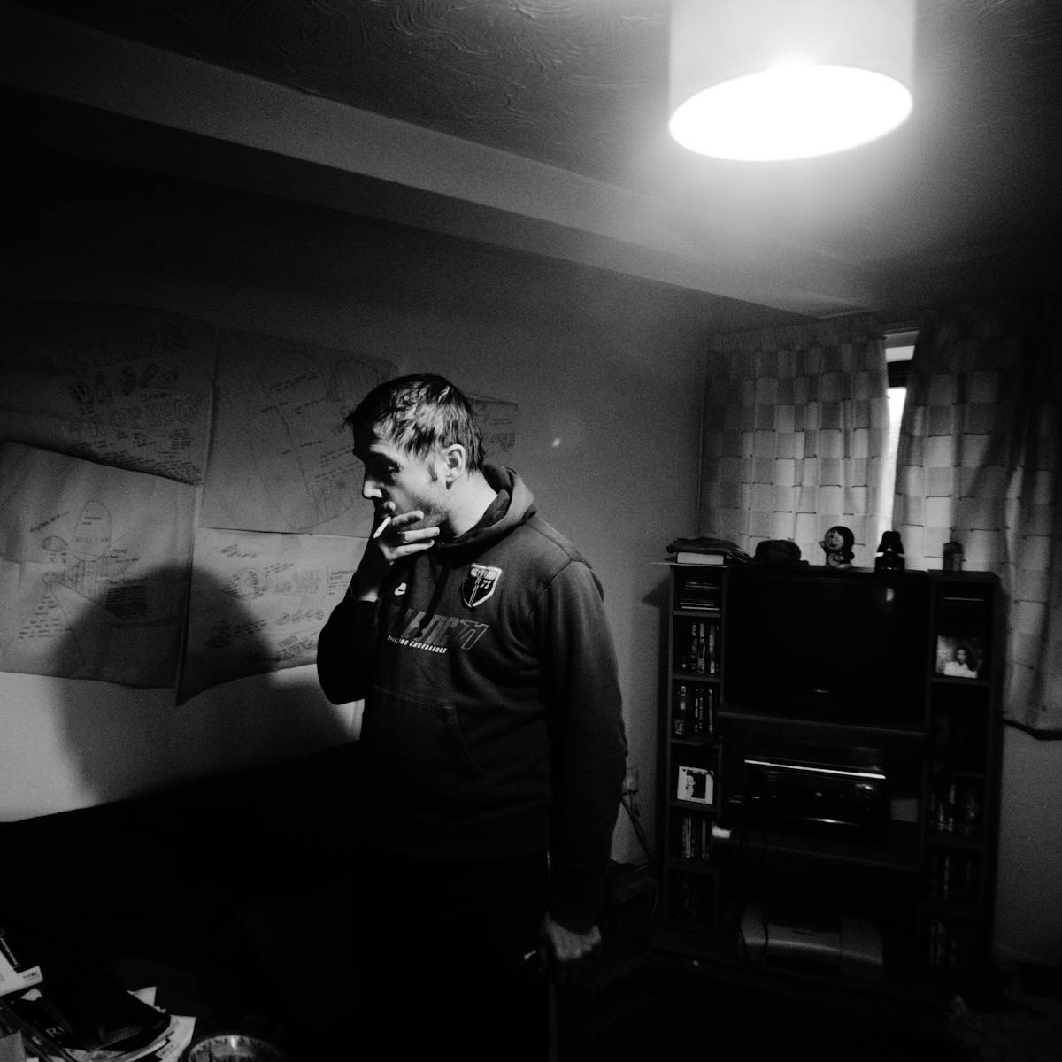 Black and white photograph showing a man standing in the middle of a living room holding a cigarette to his mouth. The room is lit by a single ceiling light, the curtains are drawn across the window. On the wall behind the man sheets of paper are stuck to the wall.