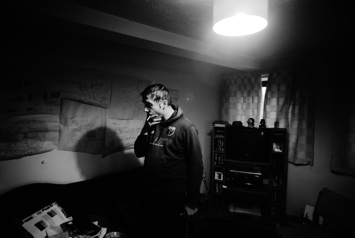 Black and white photograph showing a man standing in the middle of a living room holding a cigarette to his mouth. The room is lit by a single ceiling light, the curtains are drawn across the window. On the wall behind the man sheets of paper are stuck to the wall.