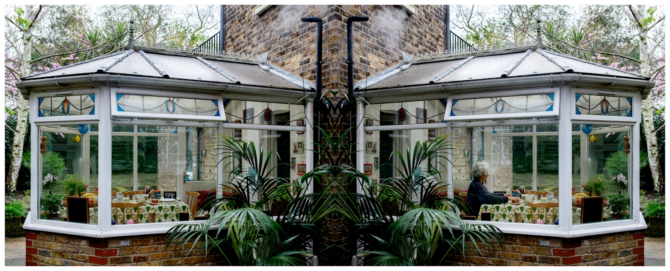 Photographic panorama showing a conservatory attached to back of a brick house. The panorama is mirrored down its vertical centre line except that a small human figure sitting at a table in the conservatory appears in the left half, but not in the right half.